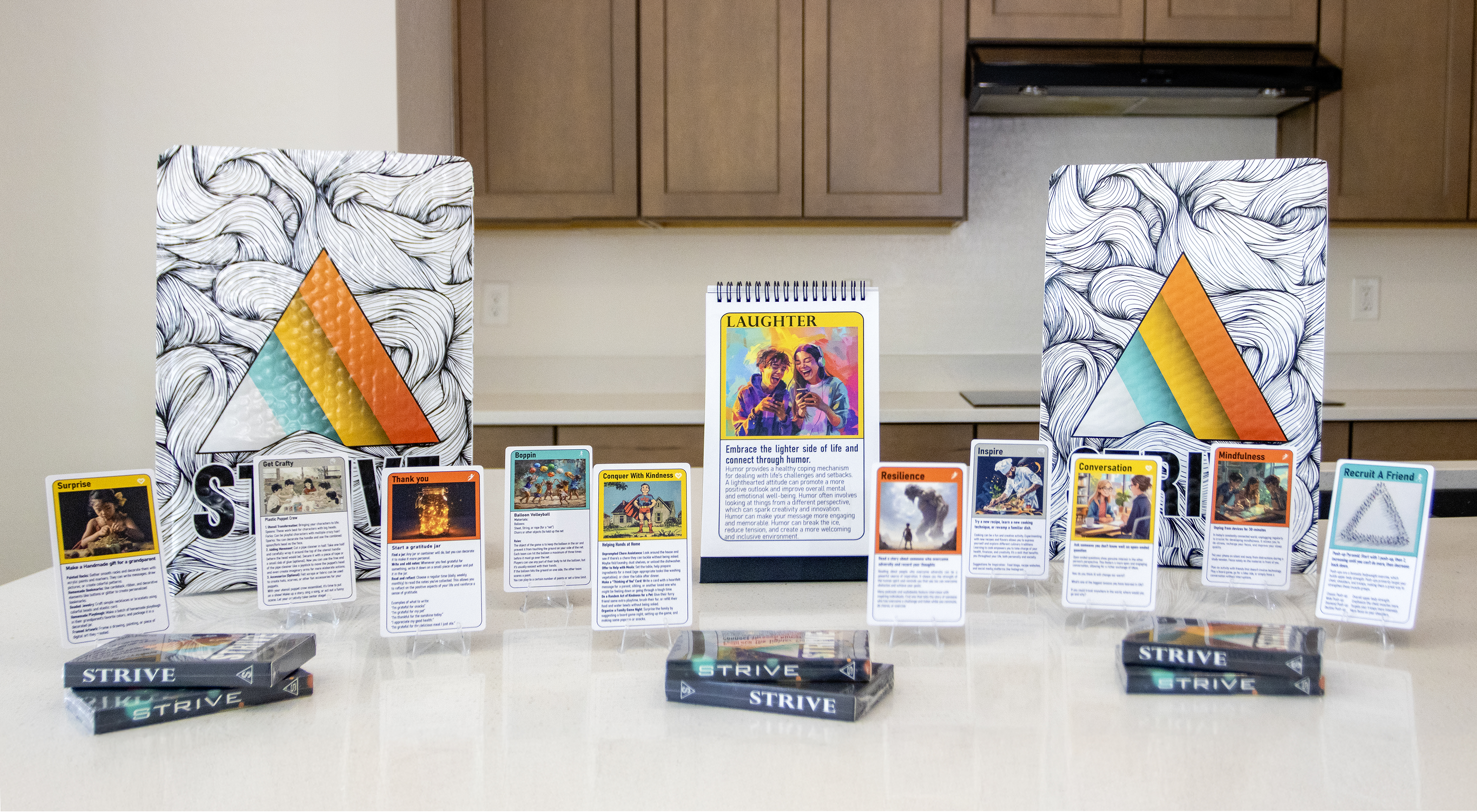 A display of the activity cards for children, activity cards for personal growth and 52-week flip book. Several cards with diverse artwork are showcased on stands, alongside two open calendars and three packaged decks. The STRIVE logo is prominent on the calendars and card backs.