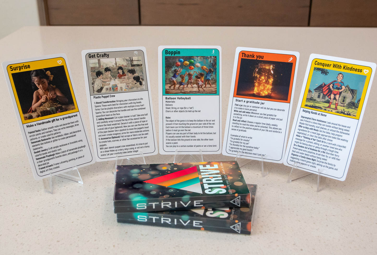 This image shows five cards from the activity cards for children deck, each with unique artwork and titles like "Get Crafty," "Boppin," and "Conquer With Kindness."  The cards are displayed on stands, and three packaged STRIVE decks are stacked in the foreground.