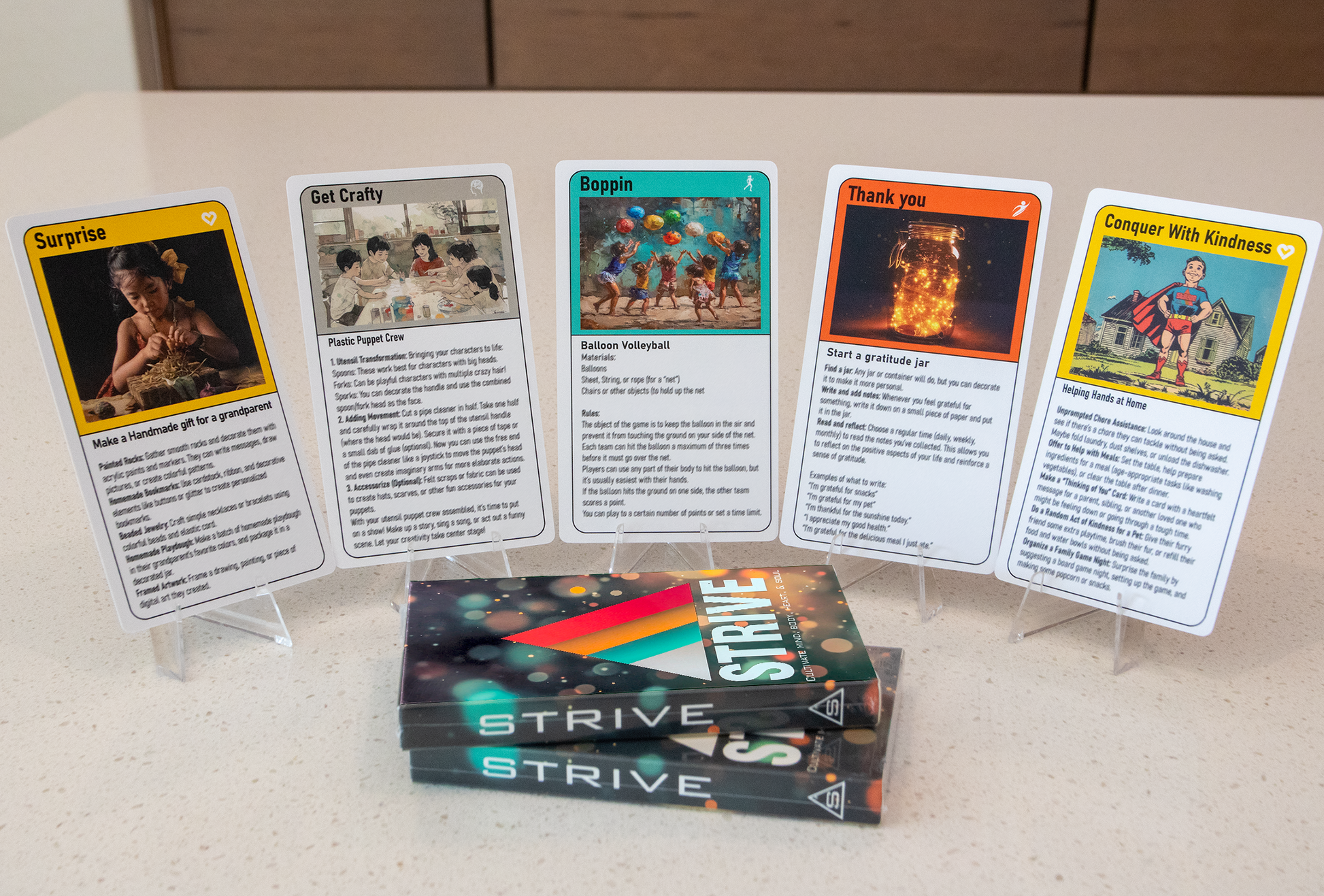 This image shows five cards from the activity cards for children deck, each with unique artwork and titles like "Get Crafty," "Boppin," and "Conquer With Kindness."  The cards are displayed on stands, and three packaged STRIVE decks are stacked in the foreground.